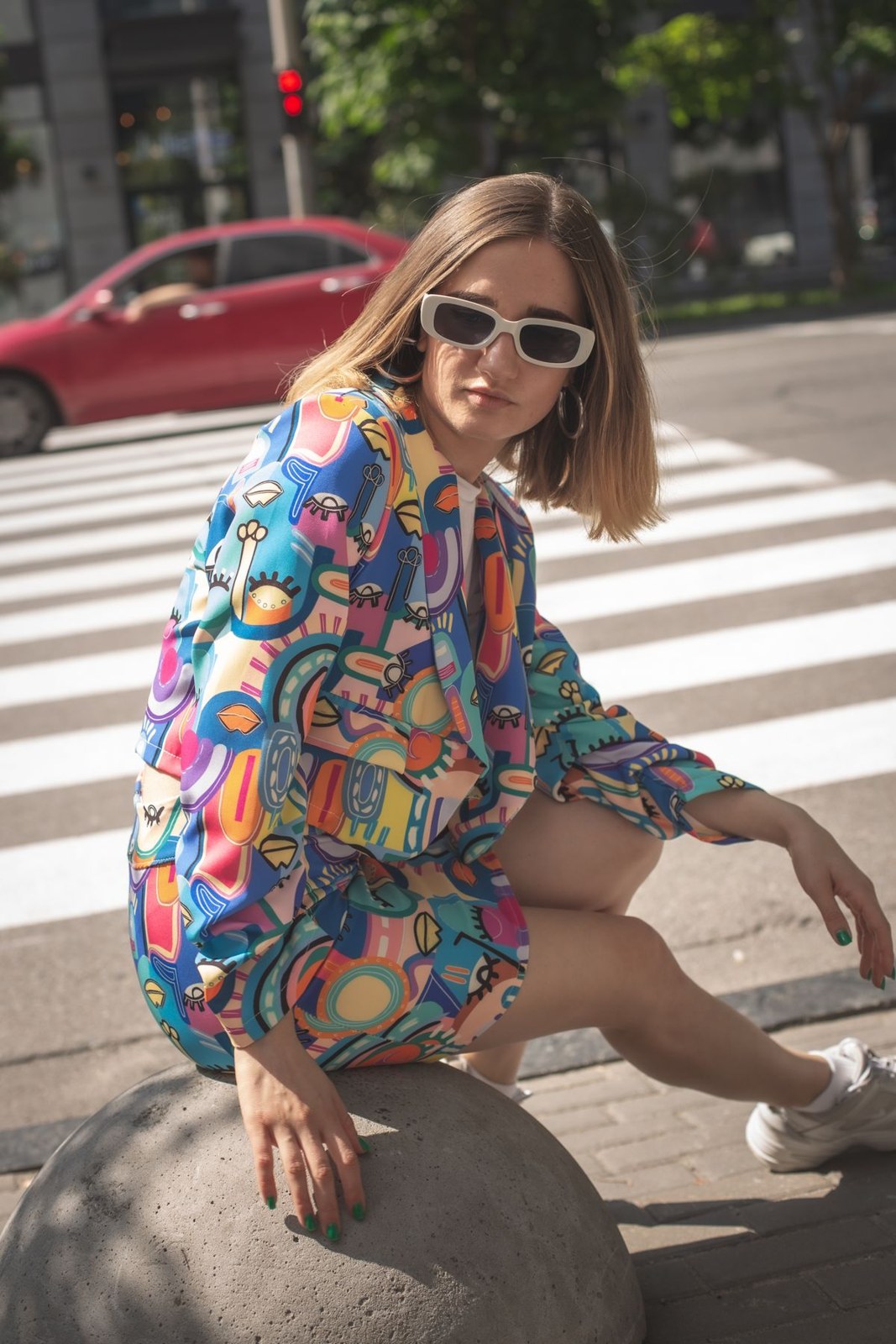 Colorful oversized printed shirt over bare legs, with pale sunglasses; seated street portrait.