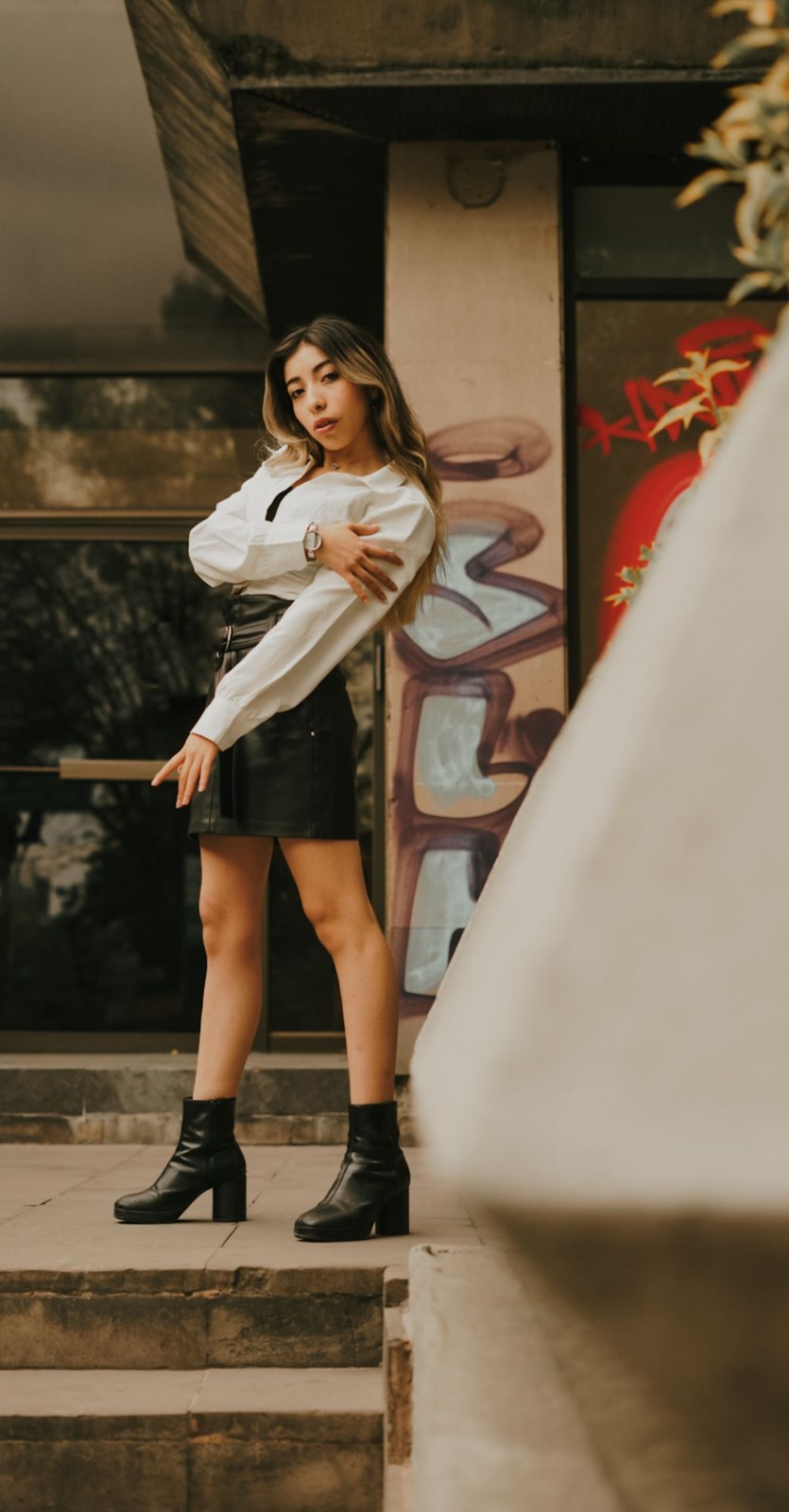White long-sleeve shirt with a black miniskirt and black ankle boots, standing on outdoor steps.