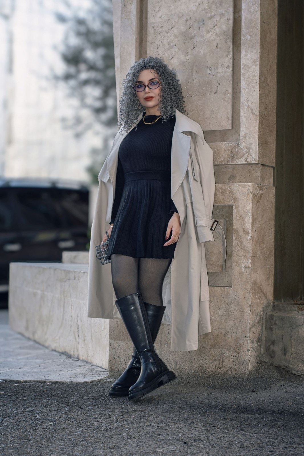 Long beige coat over a short black dress, black tights, tall black boots and sunglasses.
