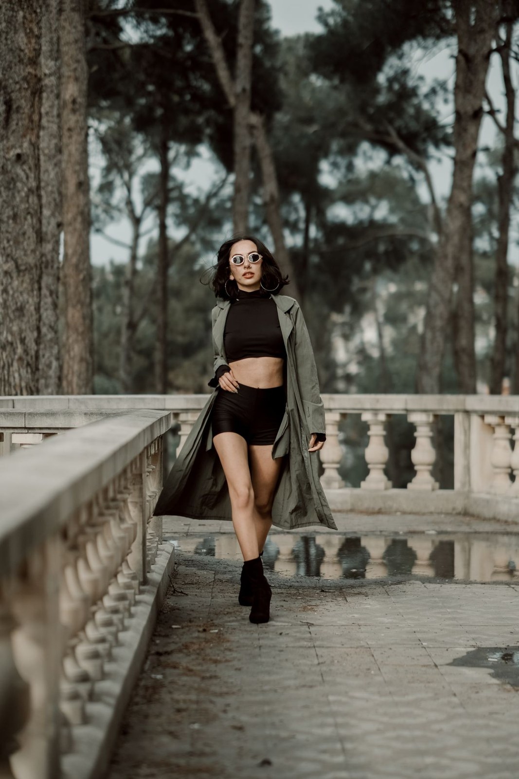 Open olive long coat over a black cropped top and black shorts, with dark ankle boots.