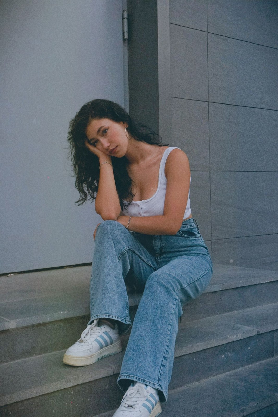 Pale tank top, loose blue jeans and white trainers, seated on steps.
