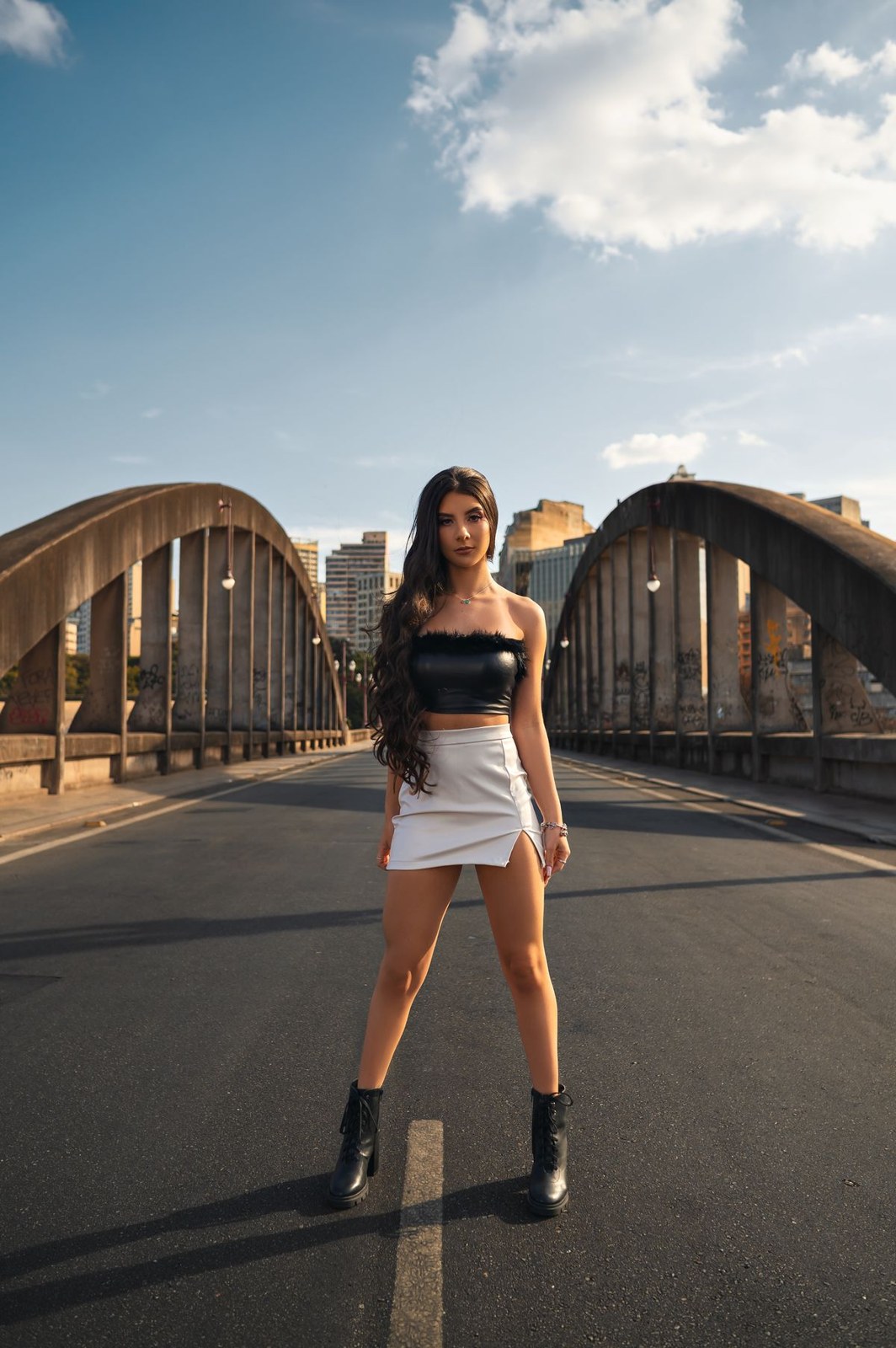 Black cropped sleeveless top, pale short skirt and black ankle boots on a bridge.