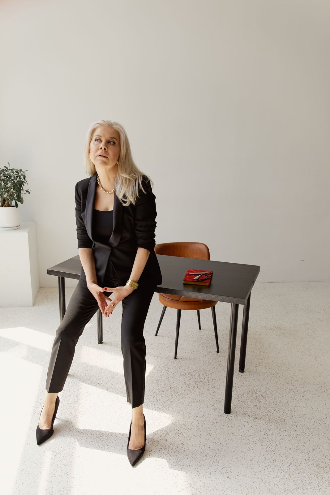 Black blazer and black top with slim dark cropped trousers and black pointed heels, seated in a bright office.