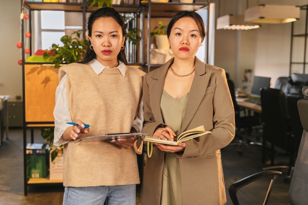 Two women in tan blazers with light tops, holding a tablet and an open book; cropped below the hips.