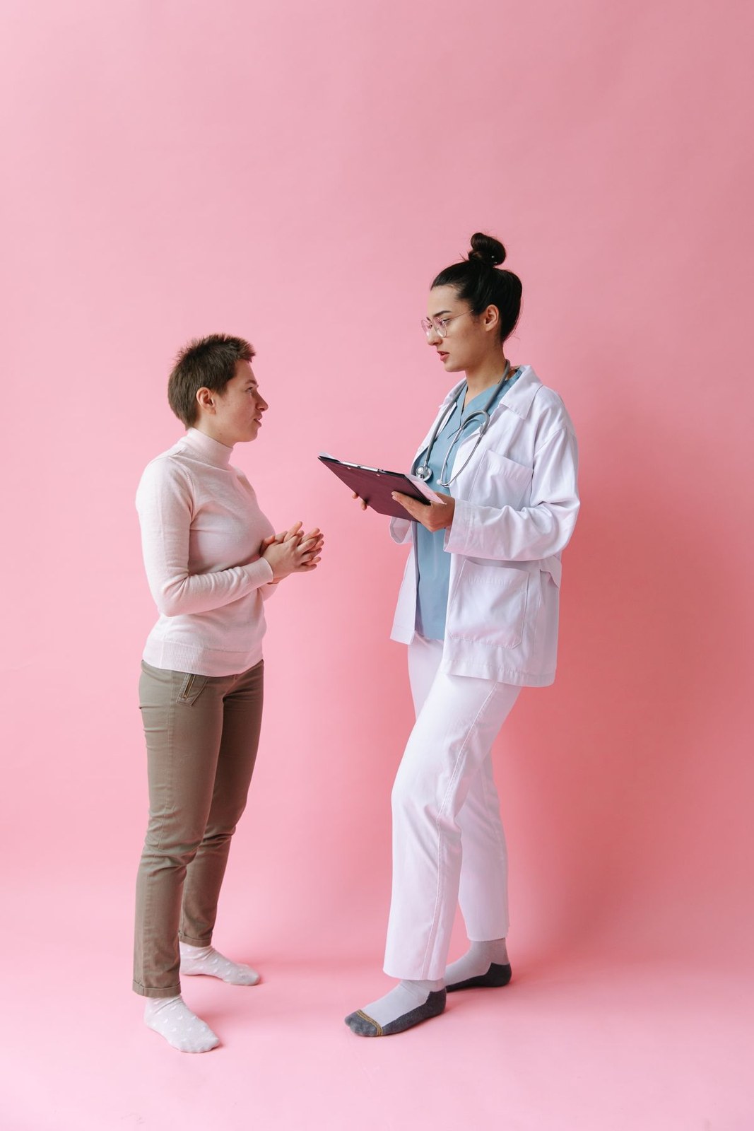 Pink sweater, tan trousers and white trainers beside a woman in a white work coat over white trousers.