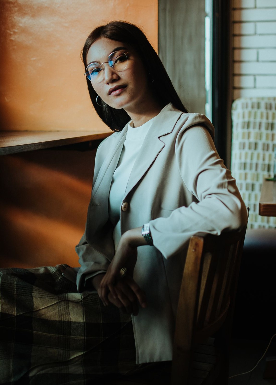 Light neutral blazer and matching trousers with glasses, seated by a bright window.