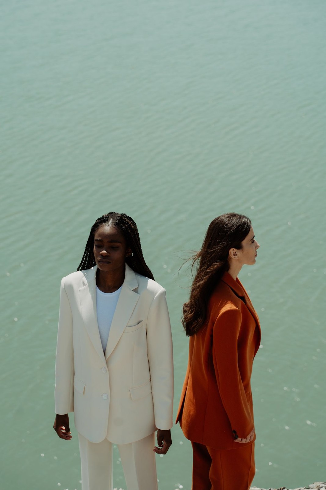 Two women in a white trouser suit and a bright orange trouser suit against a pale green wall.
