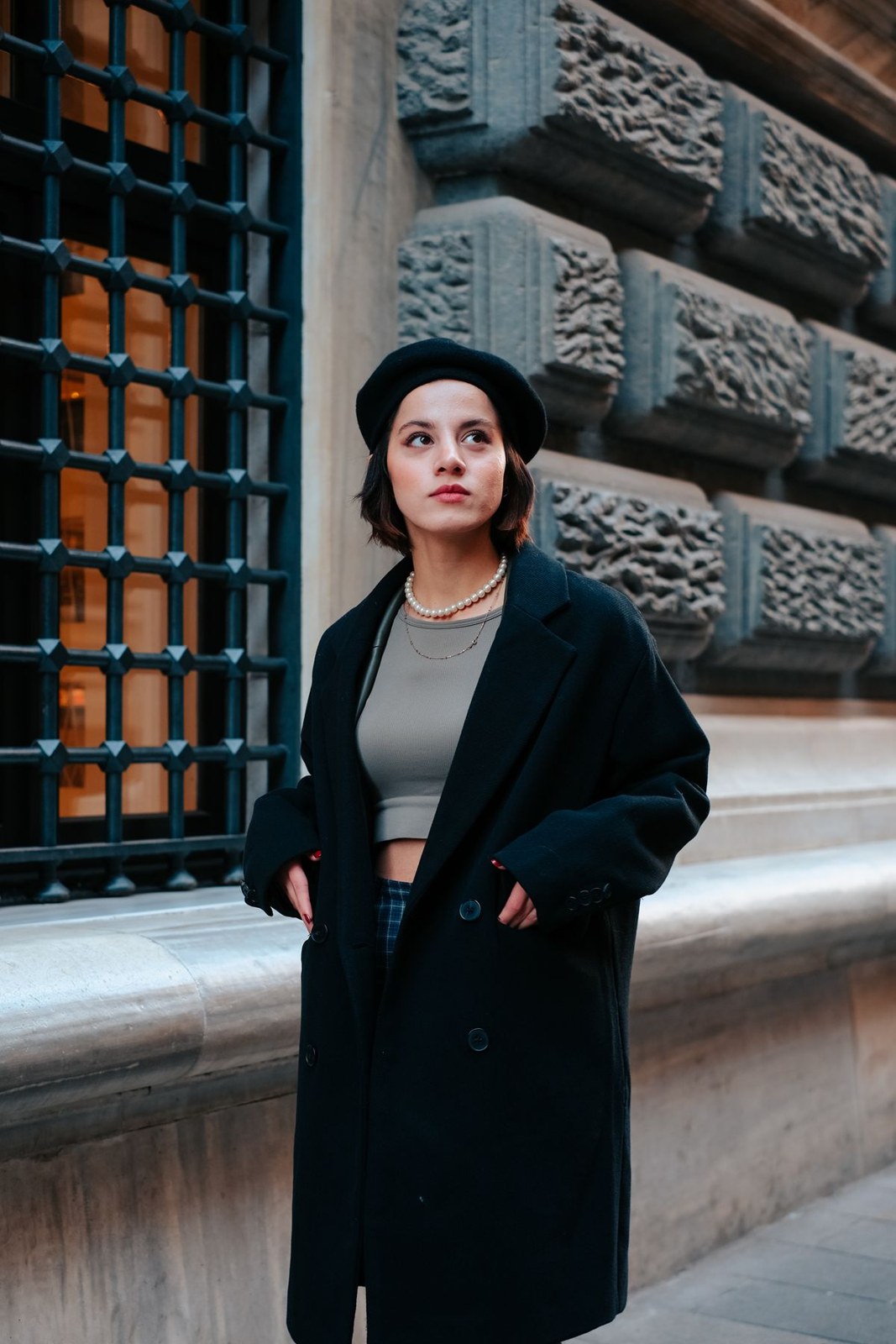 Long black coat over a grey cropped top and dark trousers, with black beret and short necklace.