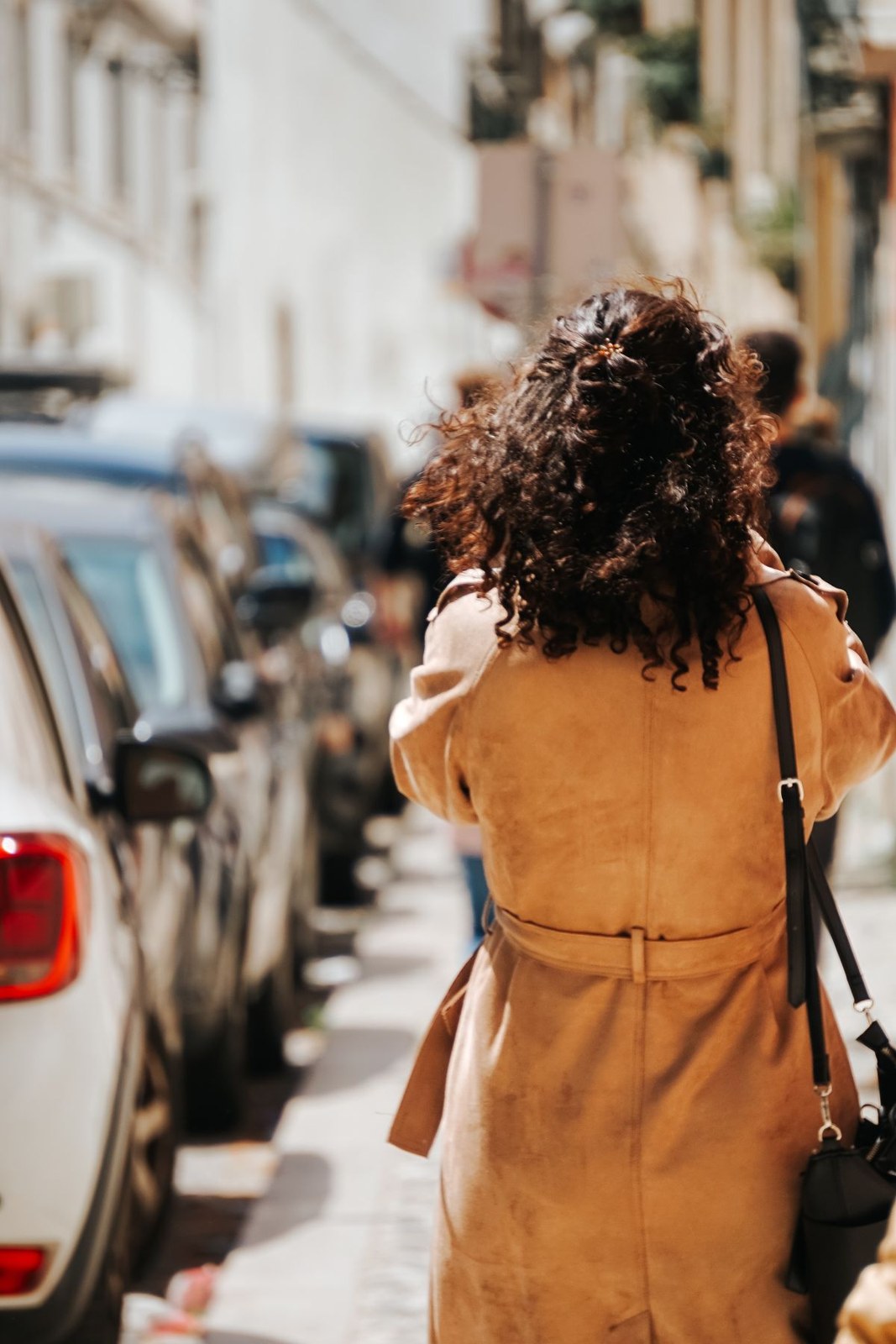 Mustard belted coat seen from the back, with a brown shoulder bag and curly hair.