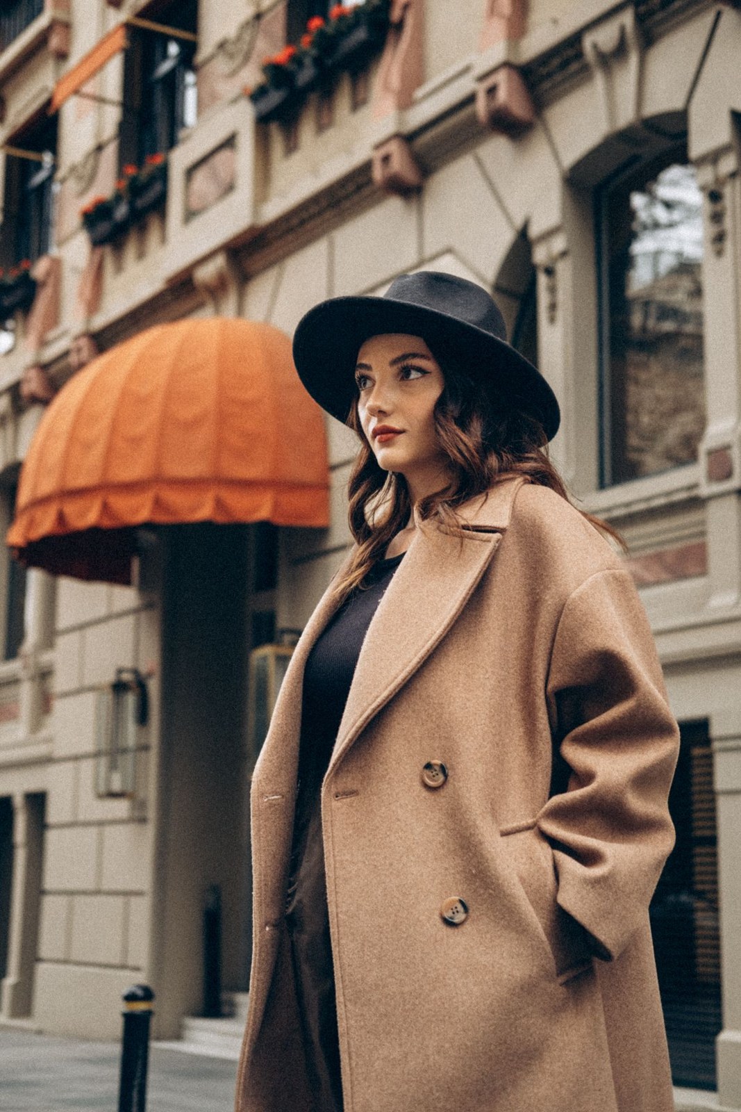 Camel coat over a black top with a wide-brimmed black hat; upper-body city portrait.