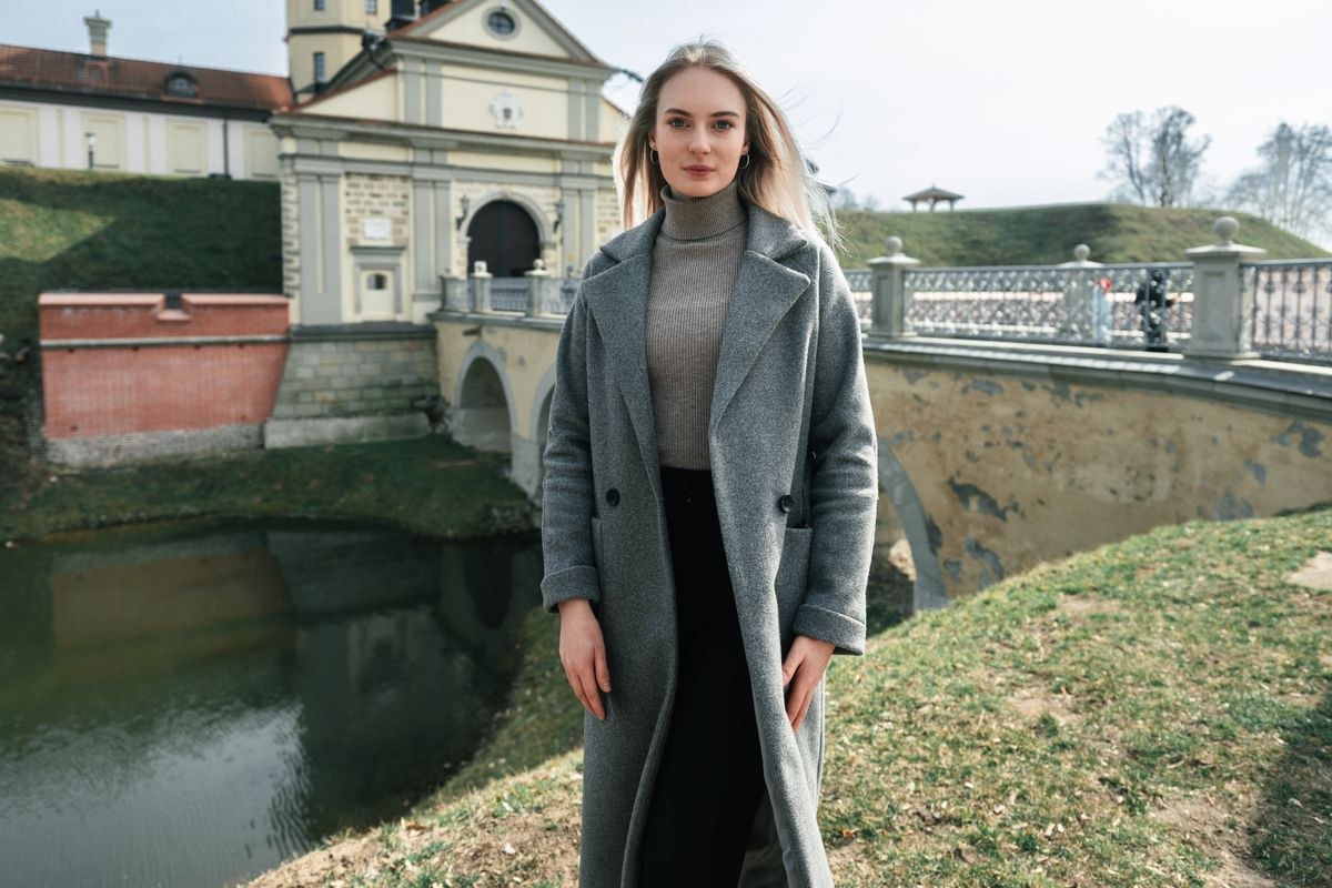 Long grey coat over a grey ribbed top and black trousers, standing by a canal.