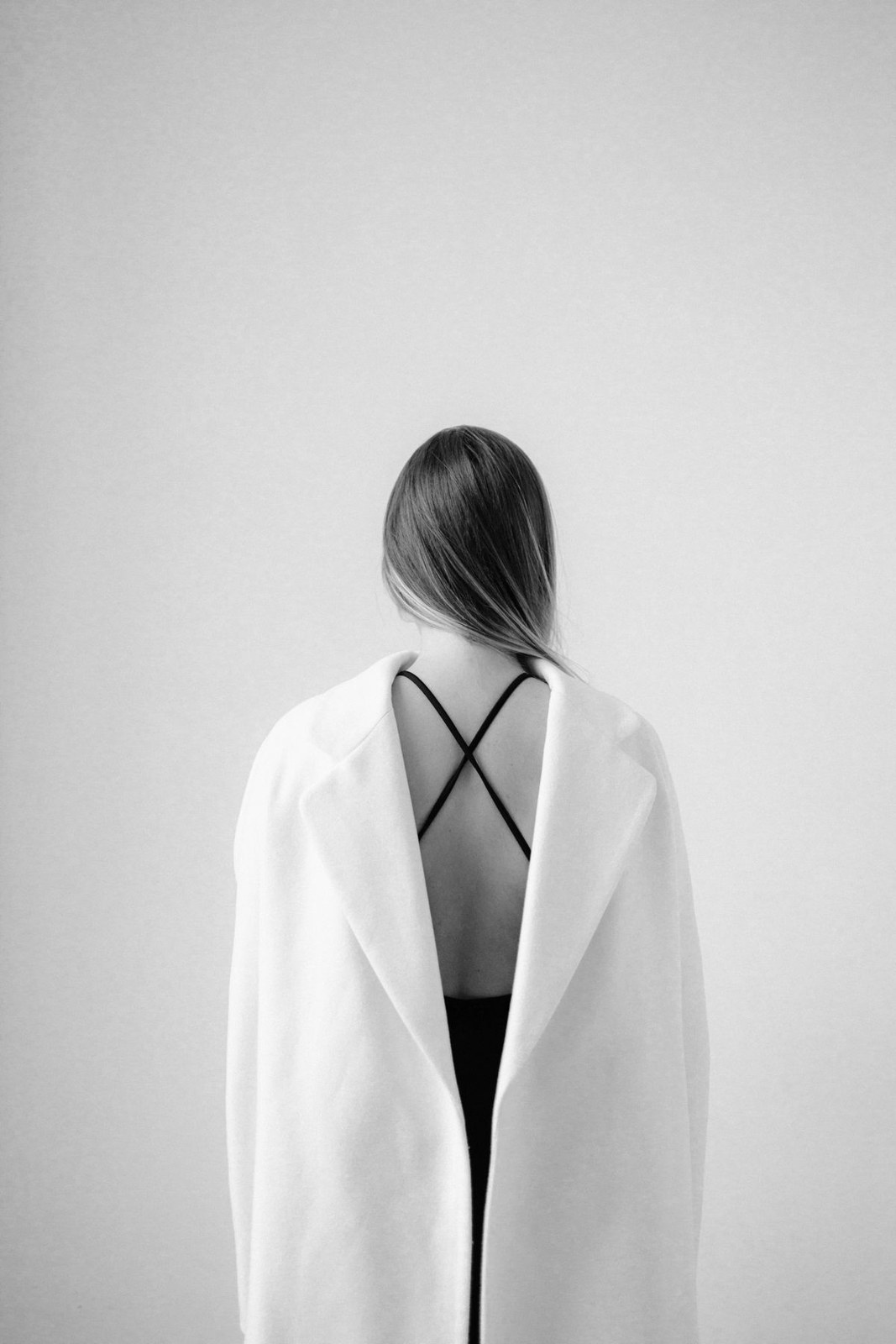 Black-and-white rear view of a light draped coat over black crossover-back straps; lower outfit not visible.
