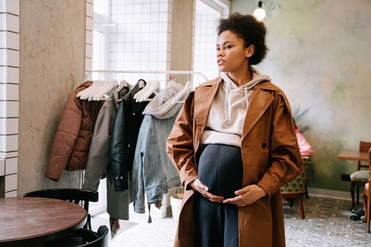 Camel coat over a light collared top and dark lower garment, photographed beside a clothing rack.