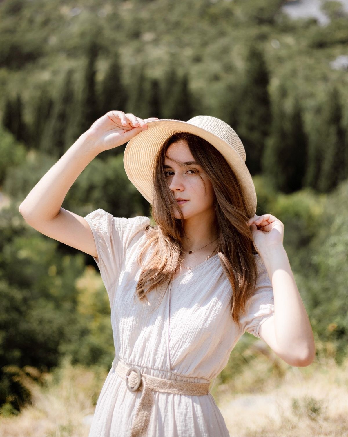 Light short-sleeve dress with a tied belt and pale wide-brim hat, cropped at the waist.