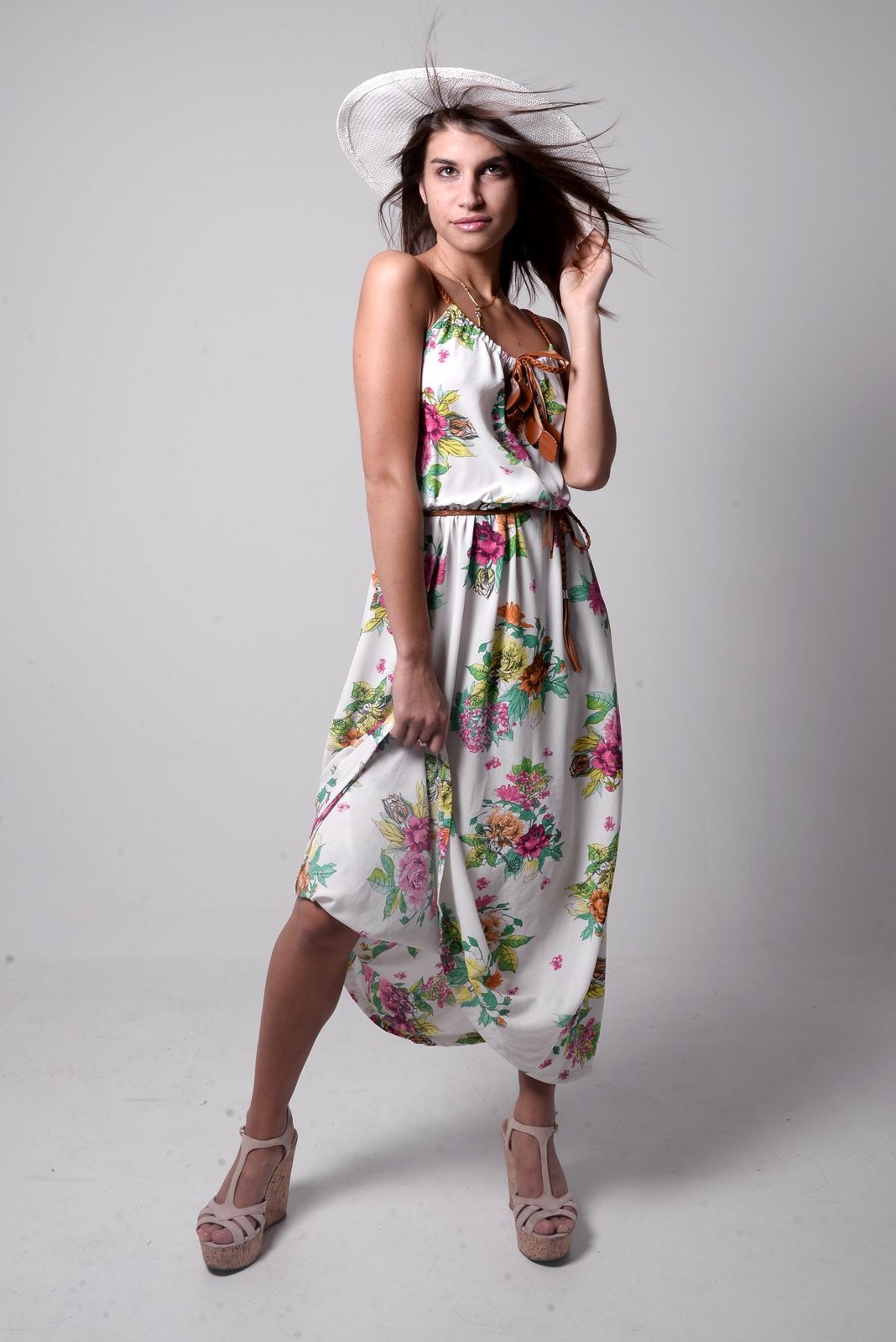 Light floral sleeveless asymmetrical dress, pale wide-brim hat and pale strappy heels in a studio.