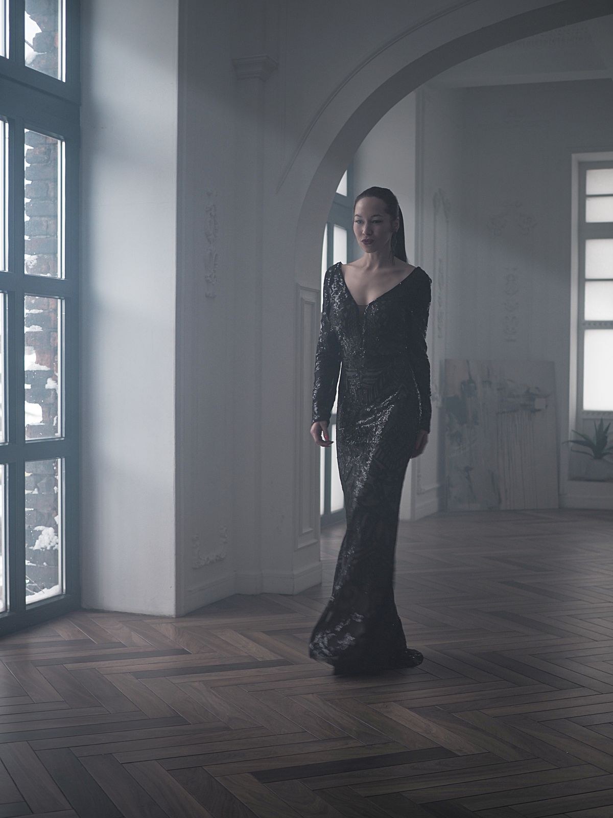 Long black V-neck gown with long sleeves in a dim room beside tall windows.