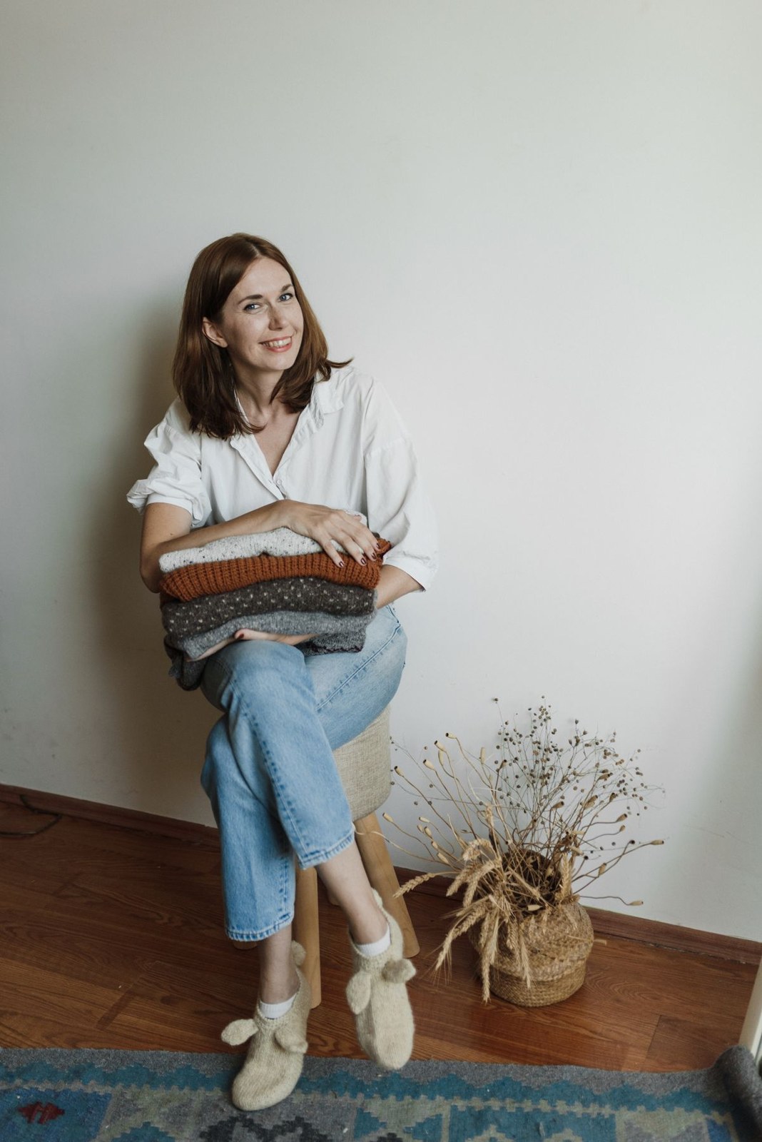 White collared blouse, cuffed light jeans and white trainers while holding folded neutral knitwear.