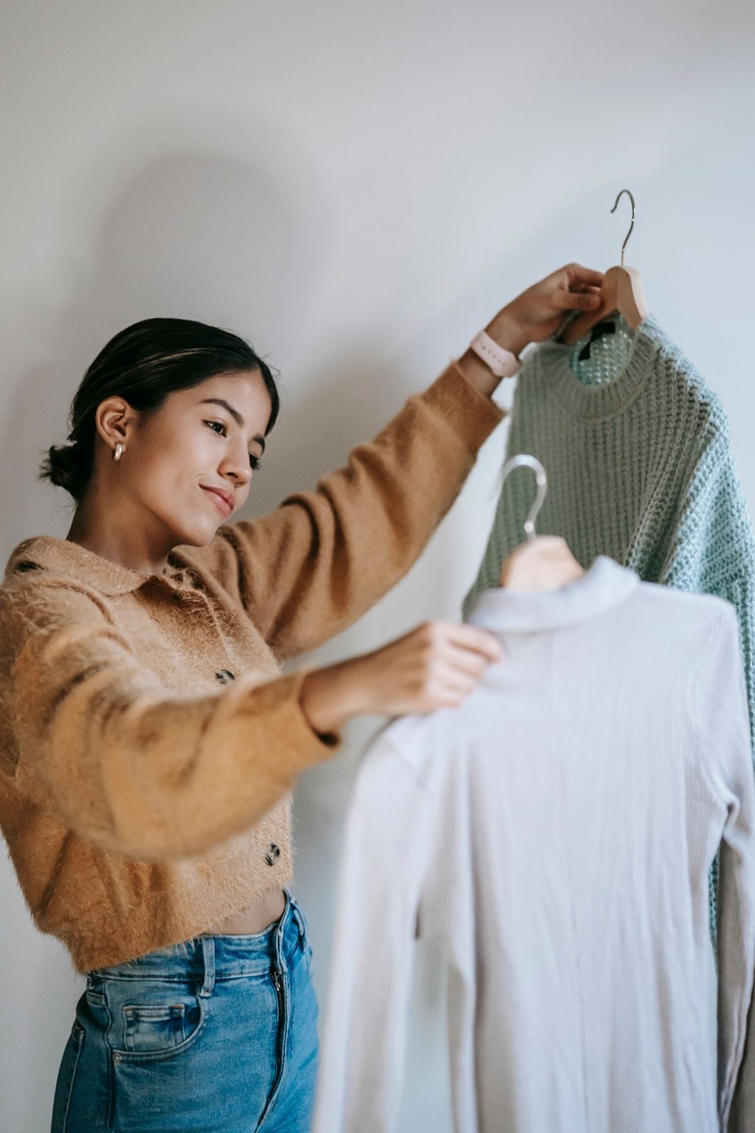 Tan buttoned cardigan and blue jeans while holding a white top and grey knit on hangers.