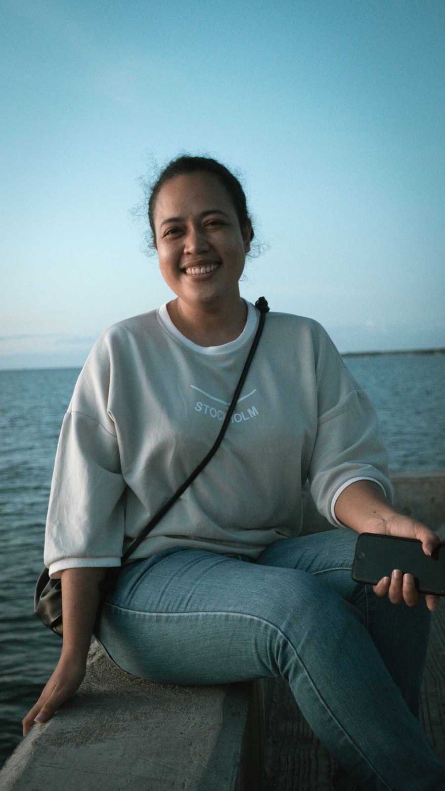 Pale-grey loose sweatshirt, cuffed blue jeans and a small black crossbody bag, seated by the sea.