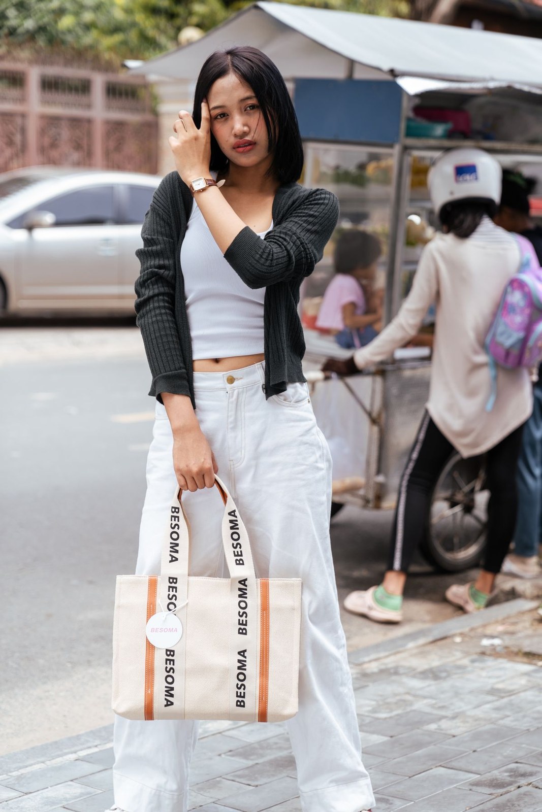 Short black cardigan over a white cropped top, long white skirt, black belt and a printed tote.