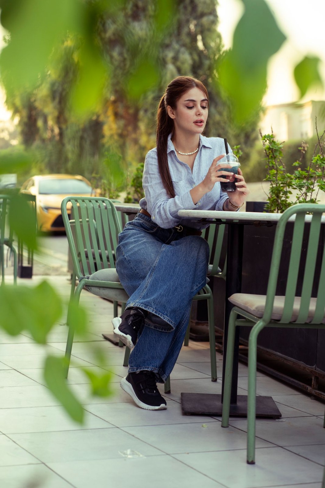 Pale collared blouse, cuffed blue jeans and black loafers while seated at an outdoor table.