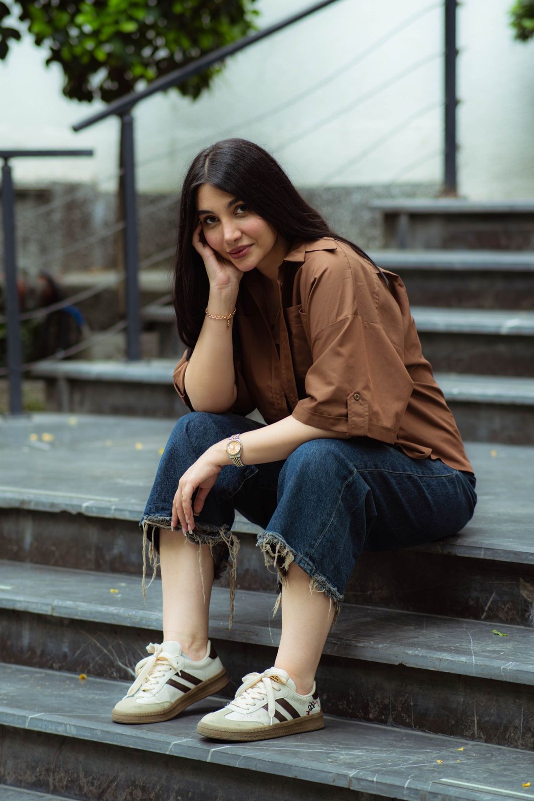 Brown long-sleeve blouse, cropped blue jeans with frayed hems and cream trainers, seated on steps.